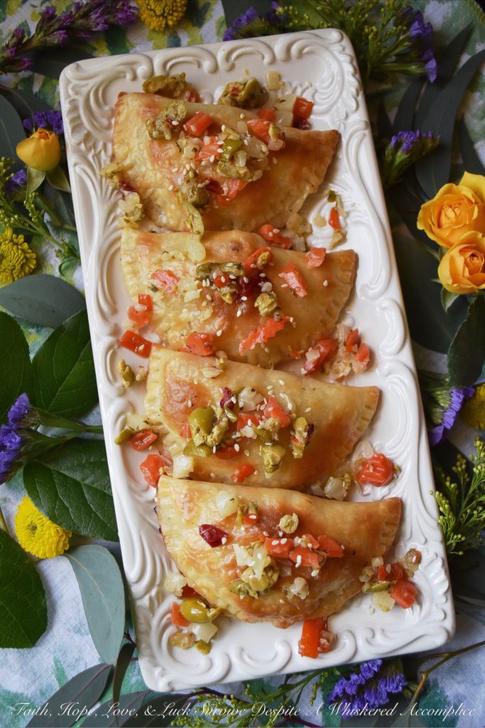 Oven-Baked Mardi Gras Muffuletta Empanadas - Looking to celebrate Mardi Gras with a unique and festive appetizer? Then these oven-baked muffuletta empanadas are just what you're looking for. Each empanada is stuffed with capicola, mortadella, salami, provolone cheese, and just the tiniest bit of Italian olive spread. After they are baked to golden brown perfection, you simply serve them topped with additional Italian olive spread and a light sprinkling of sesame seeds.
