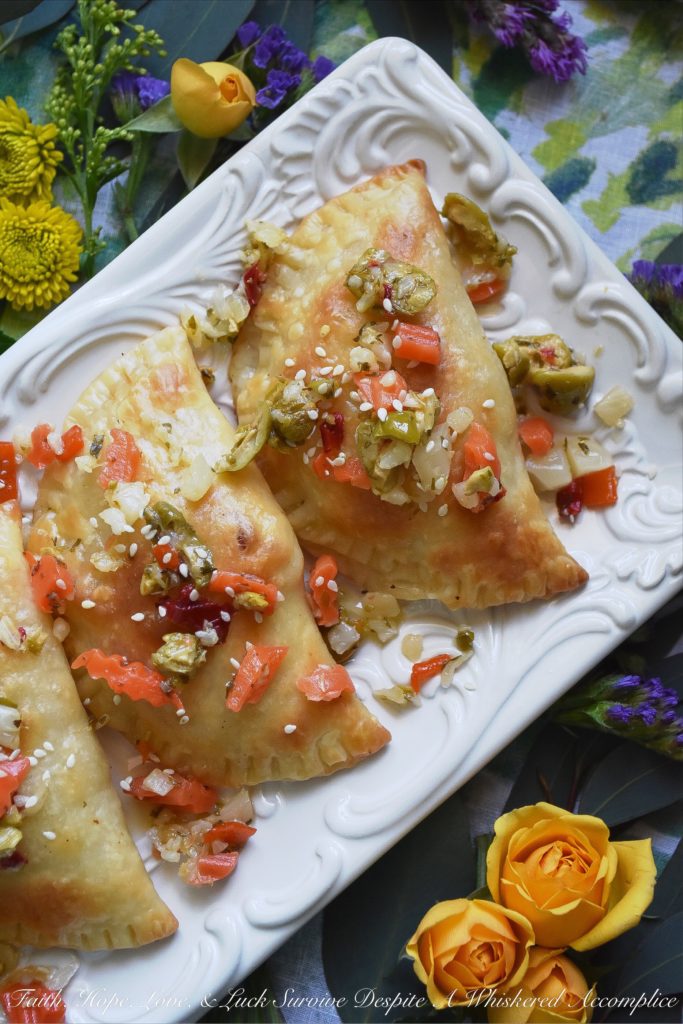 Oven-Baked Mardi Gras Muffuletta Empanadas - Looking to celebrate Mardi Gras with a unique and festive appetizer? Then these oven-baked muffuletta empanadas are just what you're looking for. Each empanada is stuffed with capicola, mortadella, salami, provolone cheese, and just the tiniest bit of Italian olive spread. After they are baked to golden brown perfection, you simply serve them topped with additional Italian olive spread and a light sprinkling of sesame seeds.