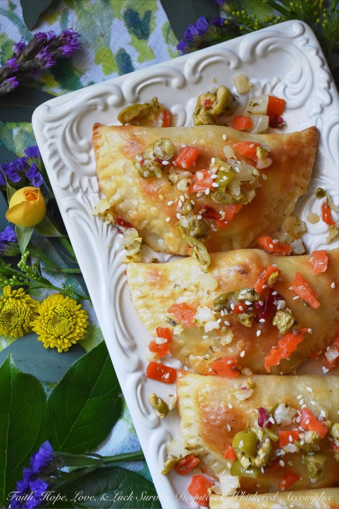 Oven-Baked Mardi Gras Muffuletta Empanadas - Looking to celebrate Mardi Gras with a unique and festive appetizer? Then these oven-baked muffuletta empanadas are just what you're looking for. Each empanada is stuffed with capicola, mortadella, salami, provolone cheese, and just the tiniest bit of Italian olive spread. After they are baked to golden brown perfection, you simply serve them topped with additional Italian olive spread and a light sprinkling of sesame seeds.